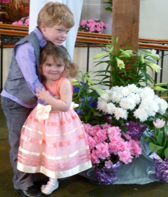 Two kids in Easter finery at the base of the flower-decked cross